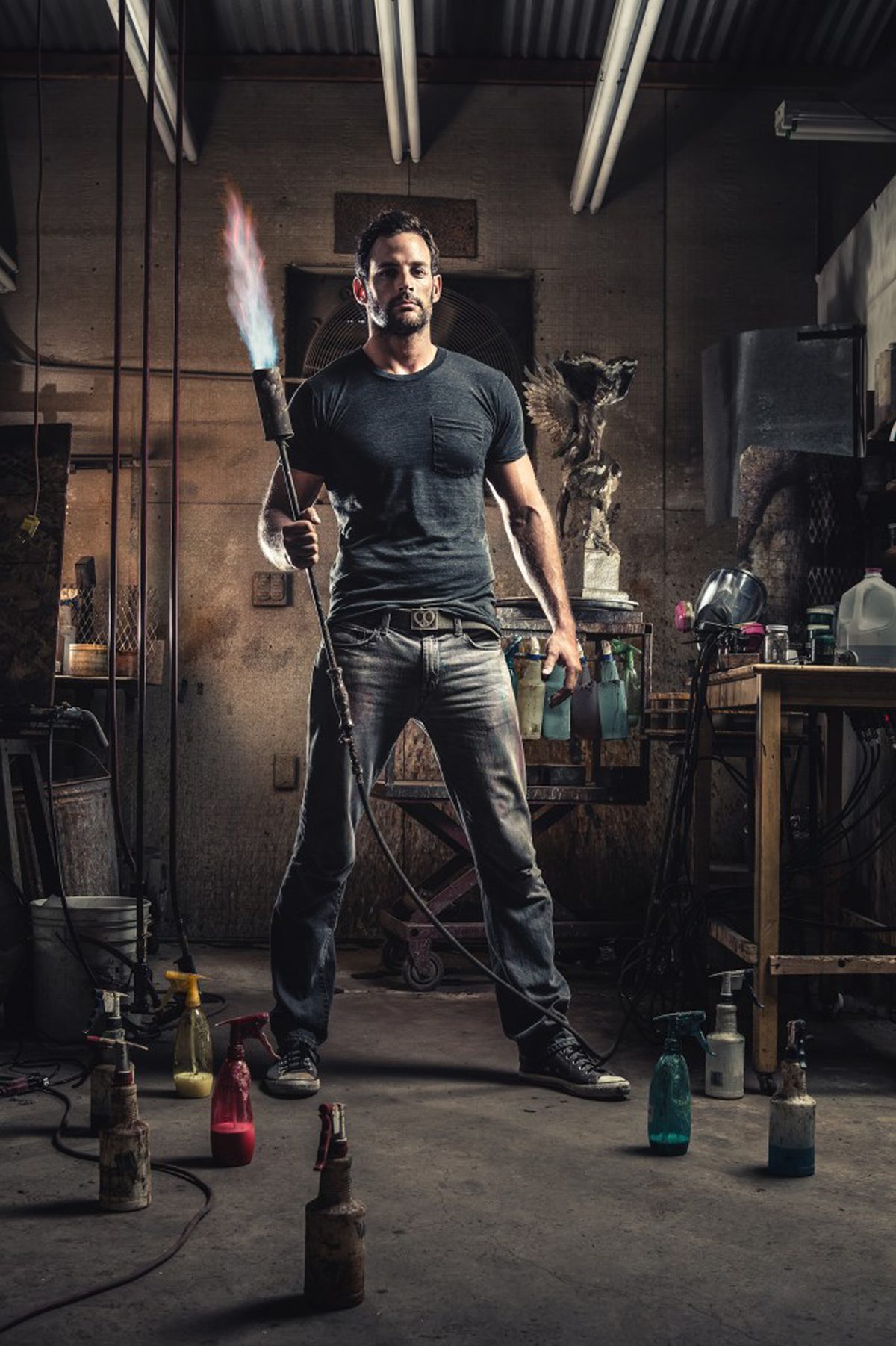 Master patineur Erik Petersen holding a lit patina torch in his studio at The Bronze Age in Prescott, Arizona, with a bronze sculpture and patina spray bottles behind him