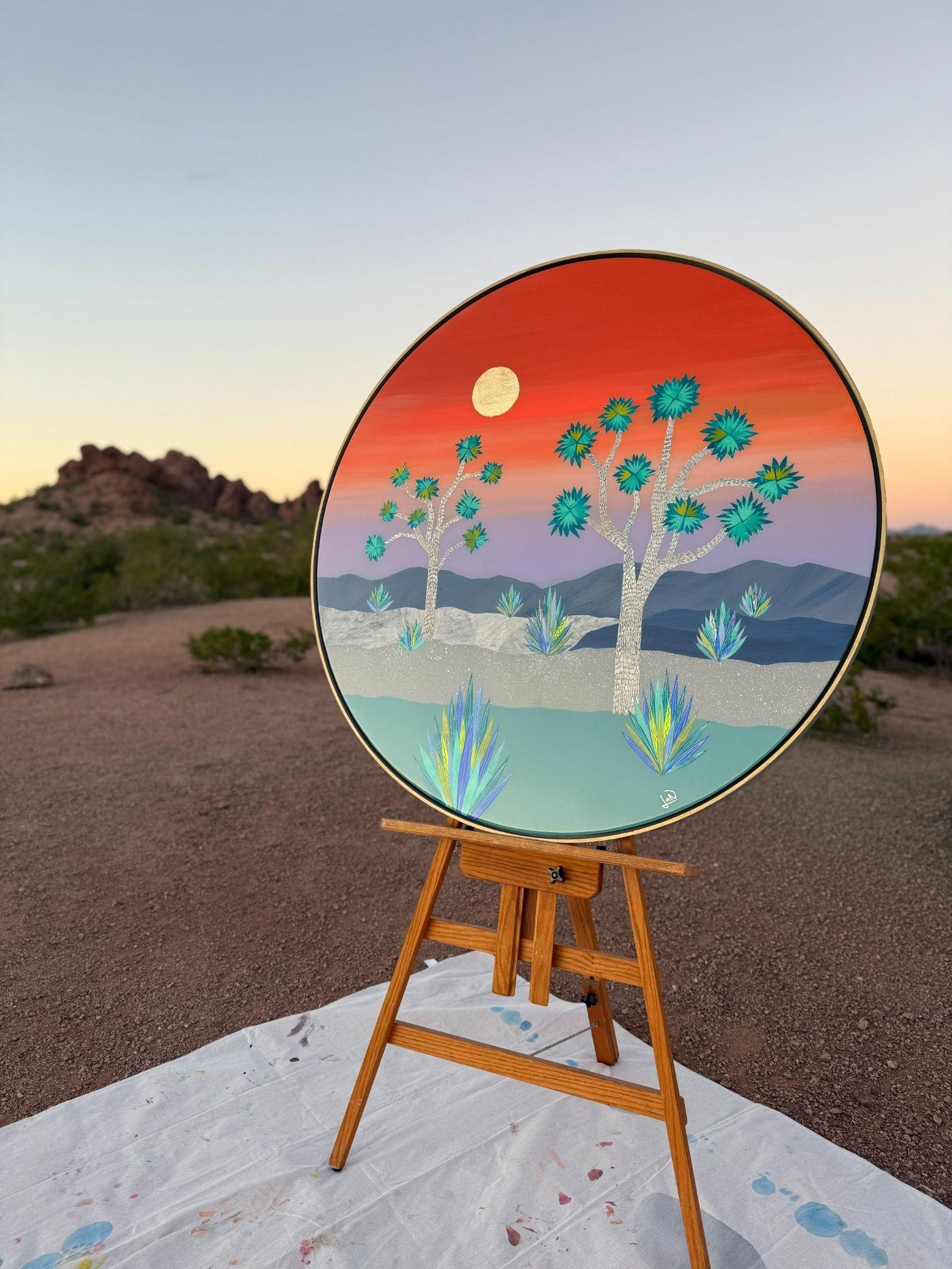 Joshua Tree Valley by Lana Nguyen, a 40-inch round paper collage under resin, on an easel in the desert at dusk